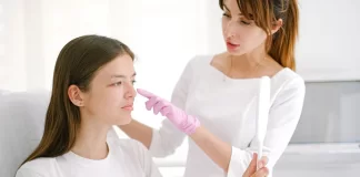Dermatolog w różowych rękawiczkach bada policzek młodej kobiety z lusterkiem.