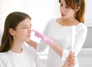 Czerwone pajączki na twarzy – jak sobie z nimi radzić? Dermatolog w różowych rękawiczkach bada policzek młodej kobiety z lusterkiem.