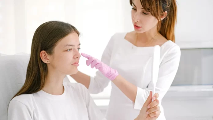 Dermatolog w różowych rękawiczkach bada policzek młodej kobiety z lusterkiem.